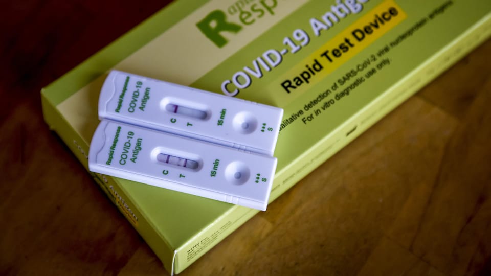 Une boîte verte contenant des tests de dépistage de COVID. Deux cassettes sont posées sur la boîte, l'une avec un résultat positif et l'autre avec un résultat négatif au test de dépistage.