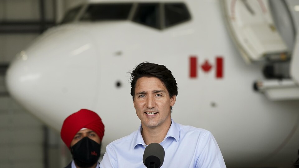 Justin Trudeau se tient devant un micro avec au fond un avion dans un hangar.