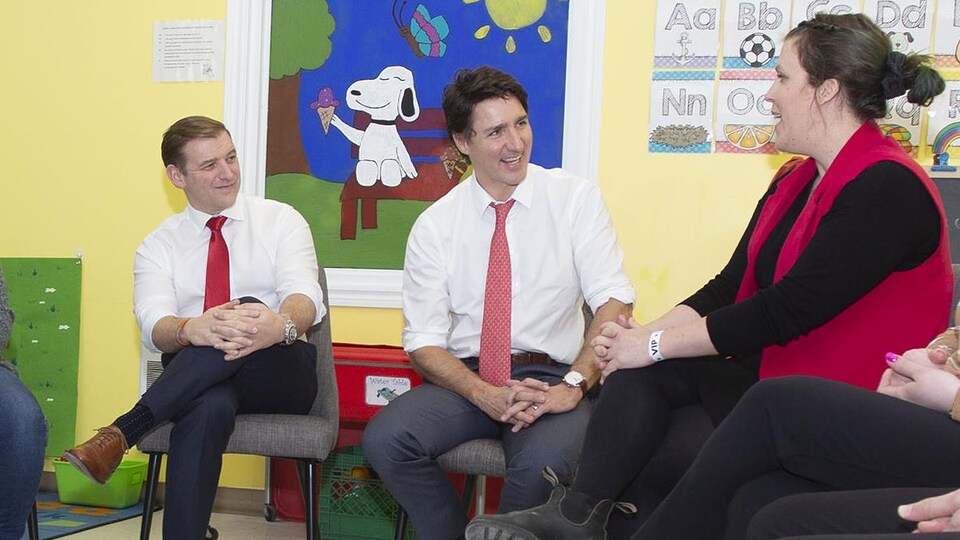 Des gens assis en rang en demi-cercle, dans une garderie. Andrew Furey et Justin Trudeau sont au milieu, en chemise blanche et cravate rouge. Ils sont assis devant un dessin de Snoopy qui tient une crème glacée.