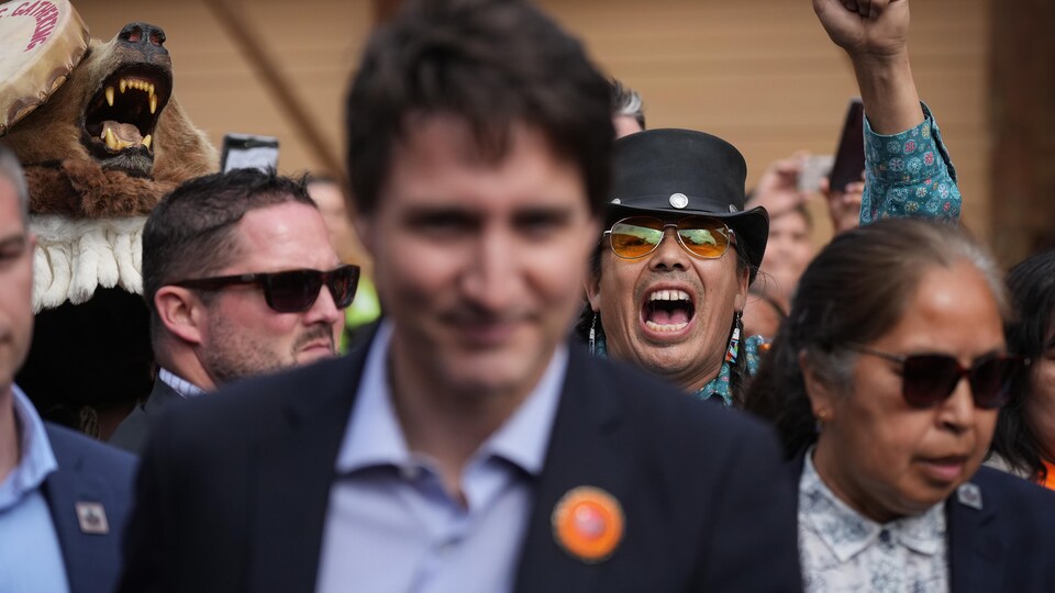Justin Trudeau passe dans une foule, derrière lui un Autochtone crie le bras levé. 