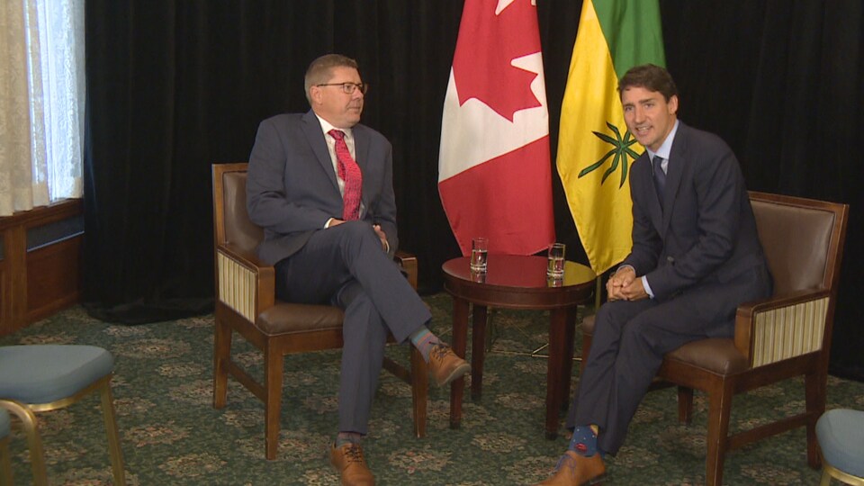 Scott Moe et Justin Trudeau assis dans des chaises devant les drapeaux de la Saskatchewan et du Canada. Scott Moe regarde Justin Trudeau avec un air grave. Justin Trudeau parle au public en souriant.