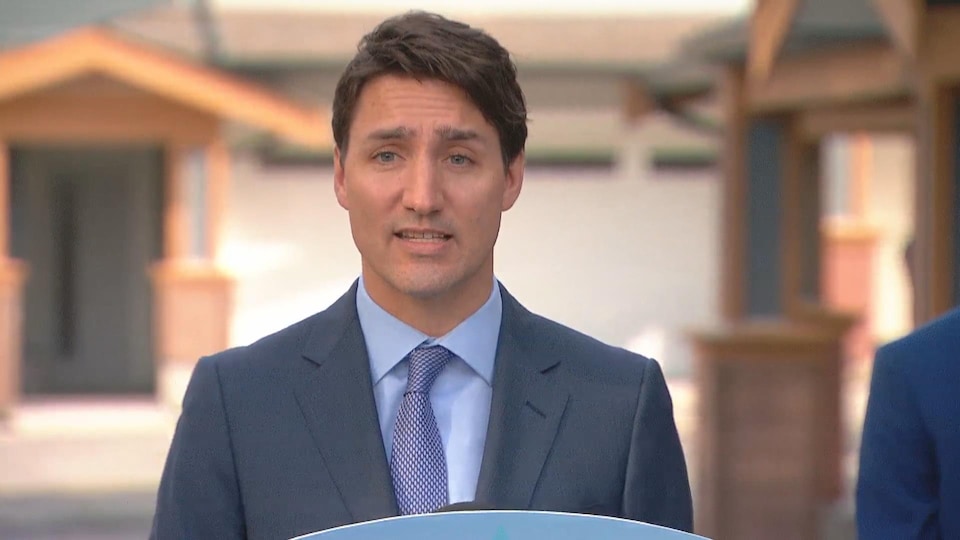 Le premier ministre vêtu d'un veston et d'une cravate regarde la caméra. On voit des maisons en arrière-plan.
