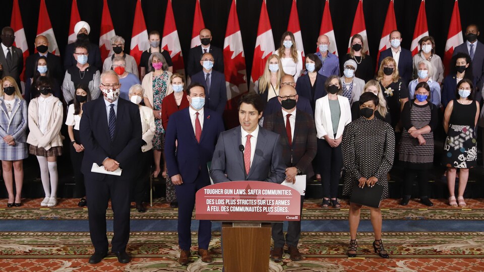 Justin Trudeau, surrounded by people wearing masks, in front of flags and in front of a lectern.