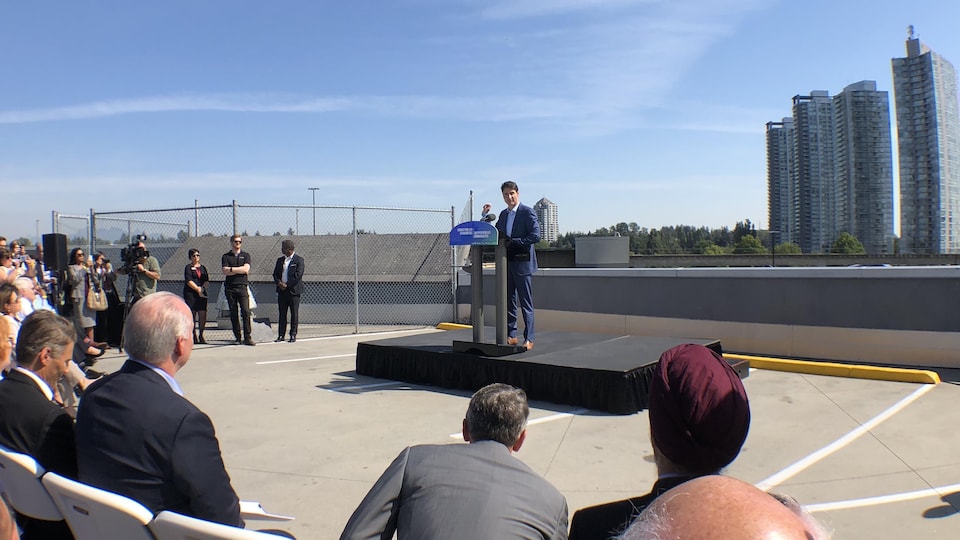 Un homme fait une présentation sur un podium entouré de spectateurs avec, en arrière-plan, une tour résidentielle.