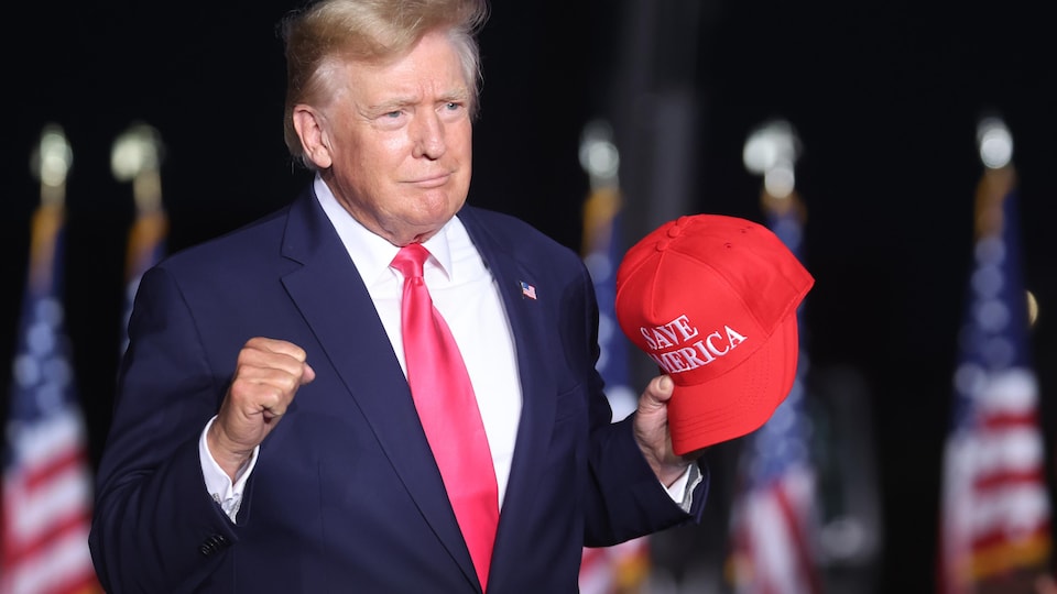 Donald Trump sur une estrade, tenant une casquette rouge.