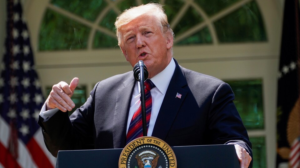 Donald Trump, la main droite levée, devant un podium, où on peut lire « président des États-Unis, dans la roseraie de la Maison-Blanche ».