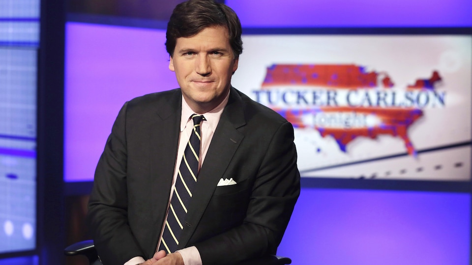 Tucker Carlson, Fox News stüdyosunda, üzerinde kendi adının yazılı olduğu bir programın başlığını gösteren ekran fotoğrafları için poz veriyor.