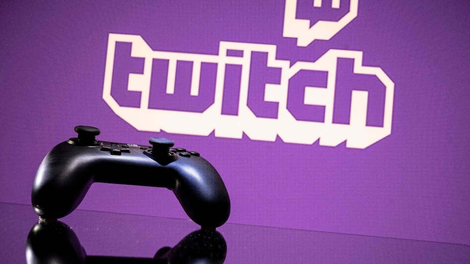Une manette de jeux vidéo est déposée sur une surface vitrée, devant un écran mauve sur lequel apparaît le mot «Twitch». 