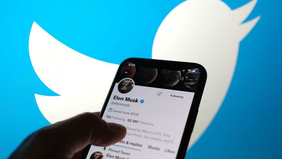 Une photo montre la main d'une personne qui tient un téléphone intelligent sur lequel on voit le compte Twitter d'Elon Musk. Derrière cela, on voit le logo de Twitter en grand format.