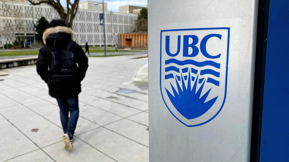 On peut voir le logo de l'université de la Colombie-Britannique au premier plan et en flou un étudiant qui marche au loin.