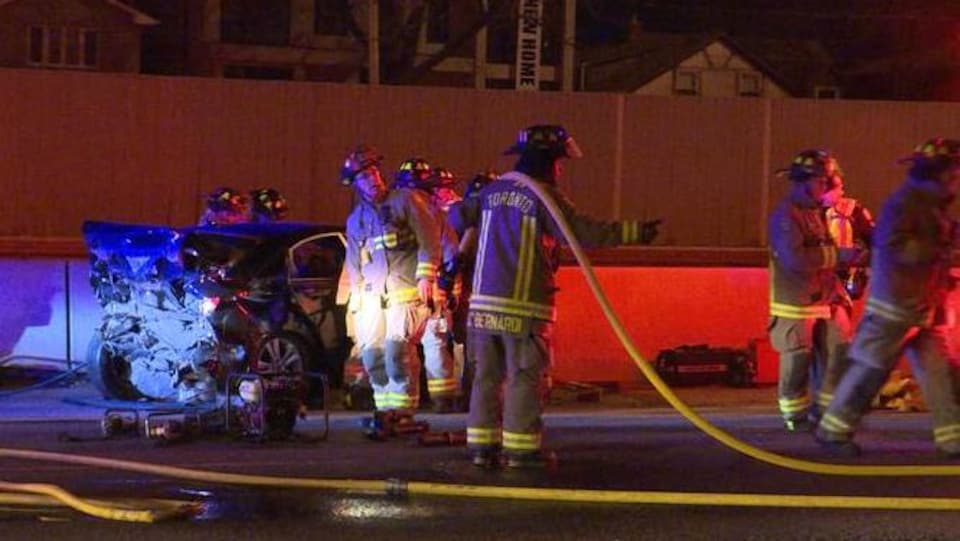 Des pompiers près d'une carcasse de voiture complètement emboutie à l'arrière.