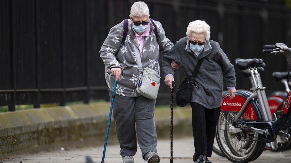 Deux personnes âgées marchent sur le trottoirs, avec chacune respectivement un masque sur le visage et une canne à main