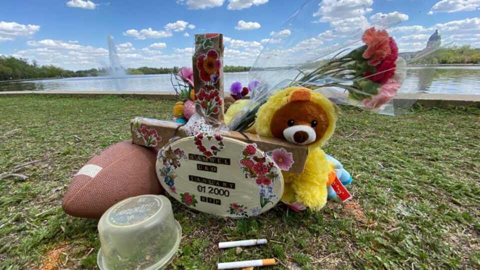 An improvised memorial with football, stuffed animals, flowers, with Wascana Lake in the background.