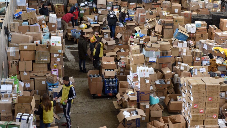 Des hommes et des femmes s'activent dans un entrepôt, au milieu de centaines de boîtes entassées. 