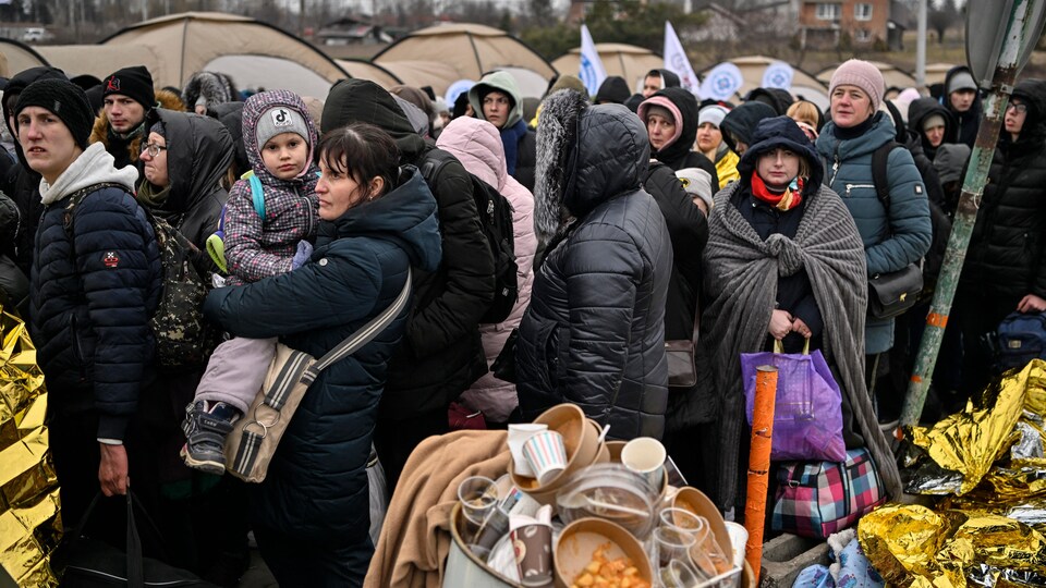 Des réfugiés, en habits et manteaux d'hiver, attendent en file. Autour d'eux, des tentes et des déchets.
