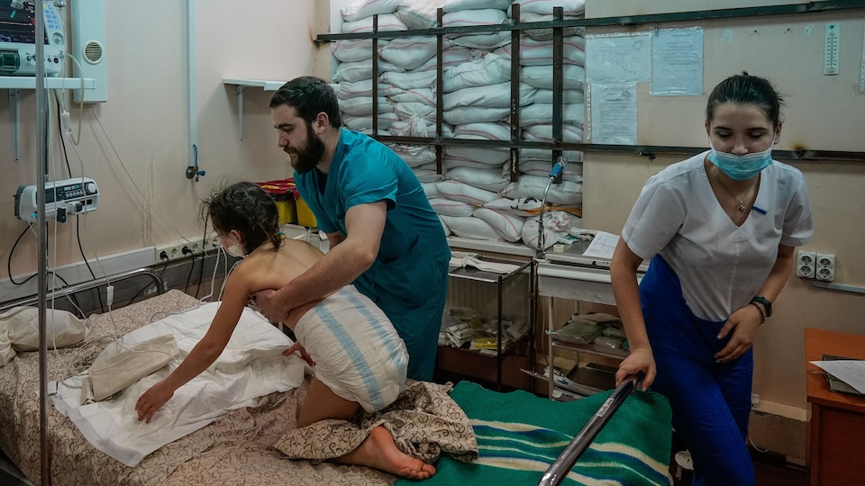 Une fillette portant seulement une couche reçoit l'aide d'un infirmier dans un lit d'hôpital. La vitre de sa chambre est protégée par des sacs de sable. 