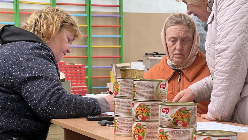 Assise à une table sur laquelle sont déposés des contenants de nourriture, Inna Michtchenko remplit des papiers à côté d'une femme plus âgée, elle-même conseillée par une autre personne.