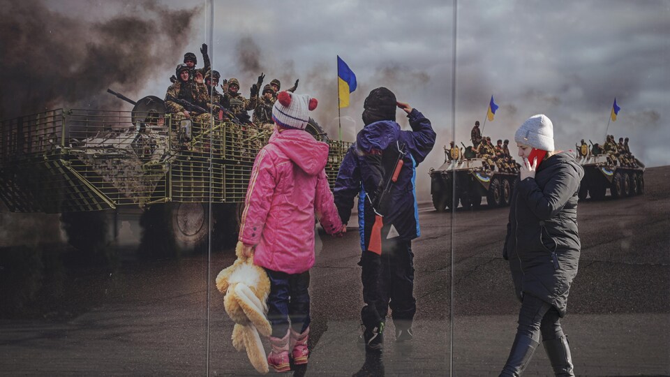 Une femme marche devant une murale montrant des enfants observant le passage de soldats ukrainiens dans des véhicules militaires.