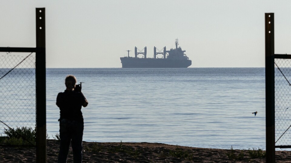 Un homme, en contre-jour, filme le départ du navire, au loin.