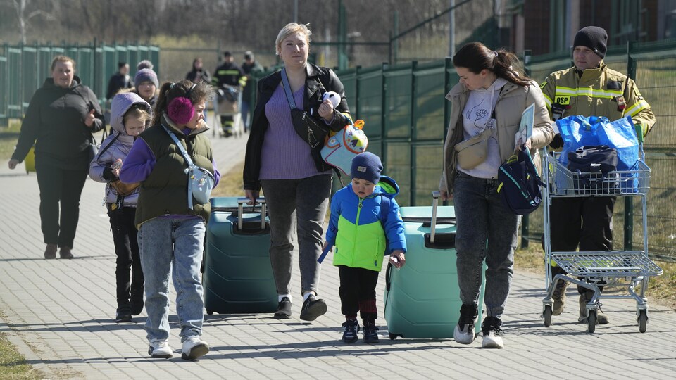 Women, children and men walk on the pavement dragging suitcases. 