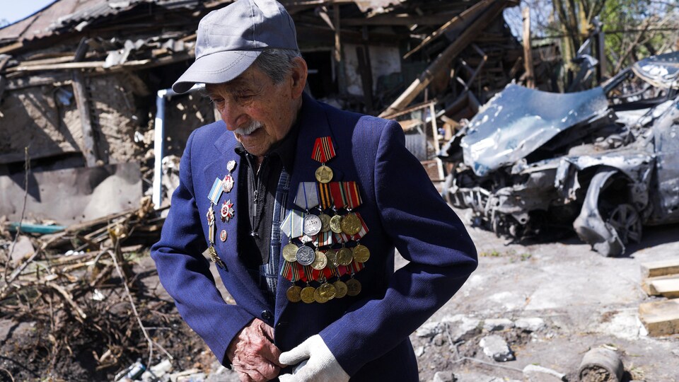 Un vieil homme boutonne un veston décoré de plusieurs médailles devant un bâtiment en ruines.
