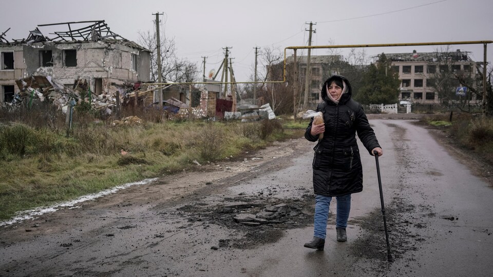 Oksana Oliinyk marche sur une route tandis qu'on voit des bâtiments détruits à l'arrière.
