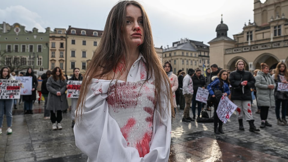Une femme portant des vêtements blancs tâchés d'un maquillage imitant le sang, au milieu d'une rue, entourée d'autres femmes portant des pancartes. 