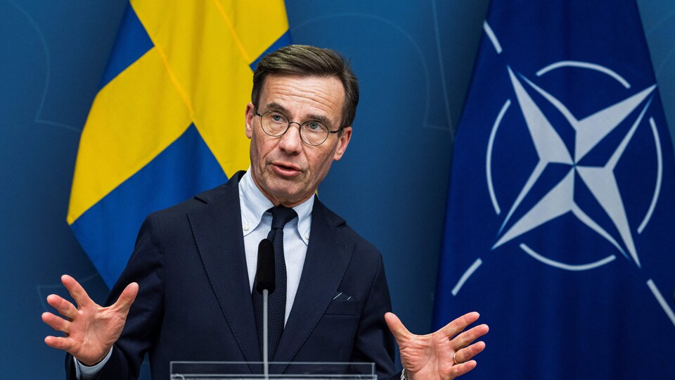 Le premier ministre de la Suède Ulf Kristersson parle dans un micro, devant des drapeaux. 