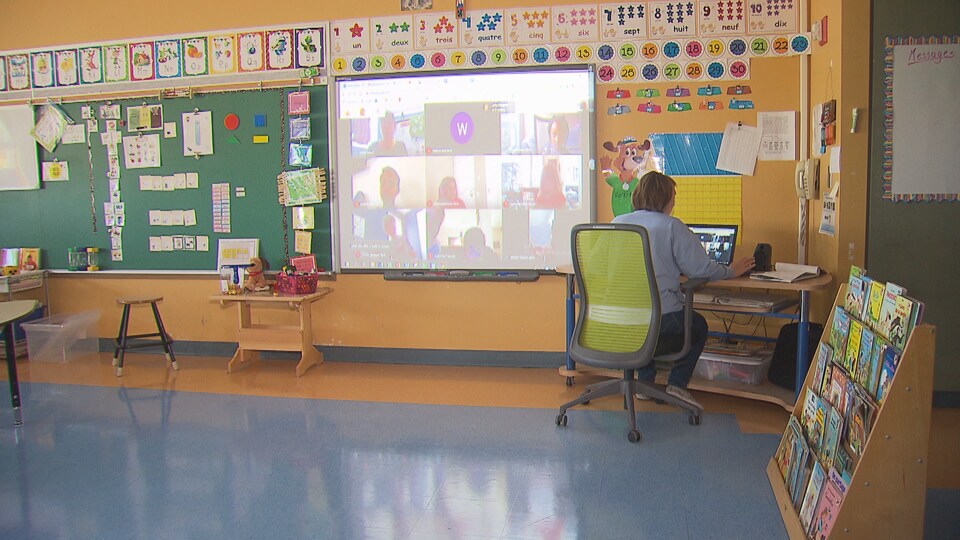 Une enseignante assise devant un ordinateur dans sa classe; au mur, un écran sur lequel est projetée une vidéoconférence, avec sa mosaïque de participants.