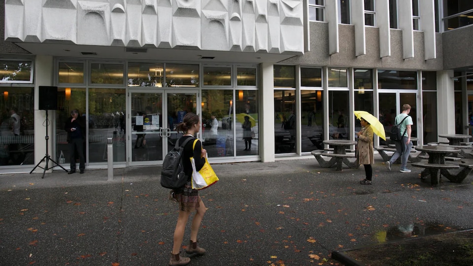 Une femme portant deux sacs marche devant un immeuble de l'Université de Victoria.