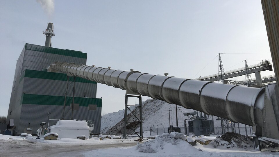 Deux bâtiments industriels sont reliés par une longue conduite de métal.