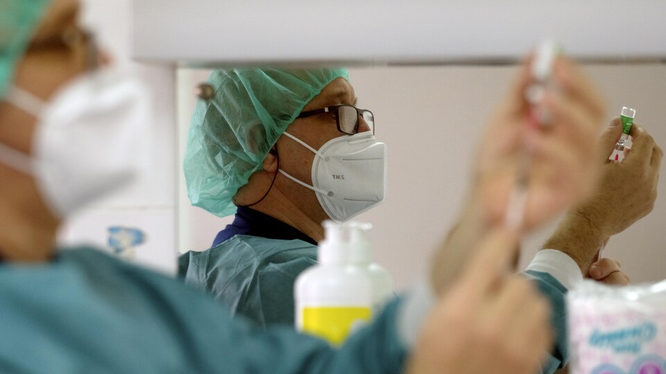 Un homme portant des équipements de protection extrait une dose de vaccin d'une fiole. On voit son reflet dans un miroir.