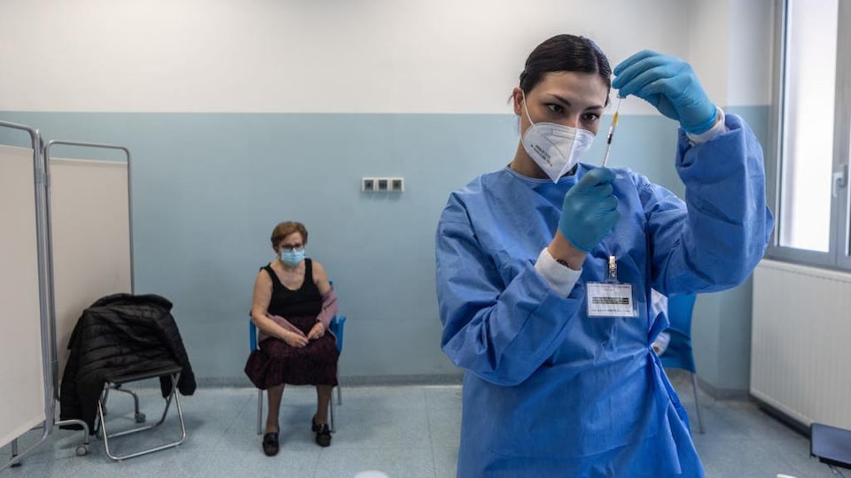 Une femme aînée est assise, avec un couvre-visage, en attendant qu'une infirmière prépare la dose de vaccin.