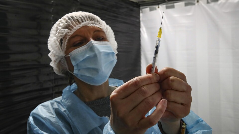Une femme portant un masque chirurgical et une blouse de plastique tient une seringue.