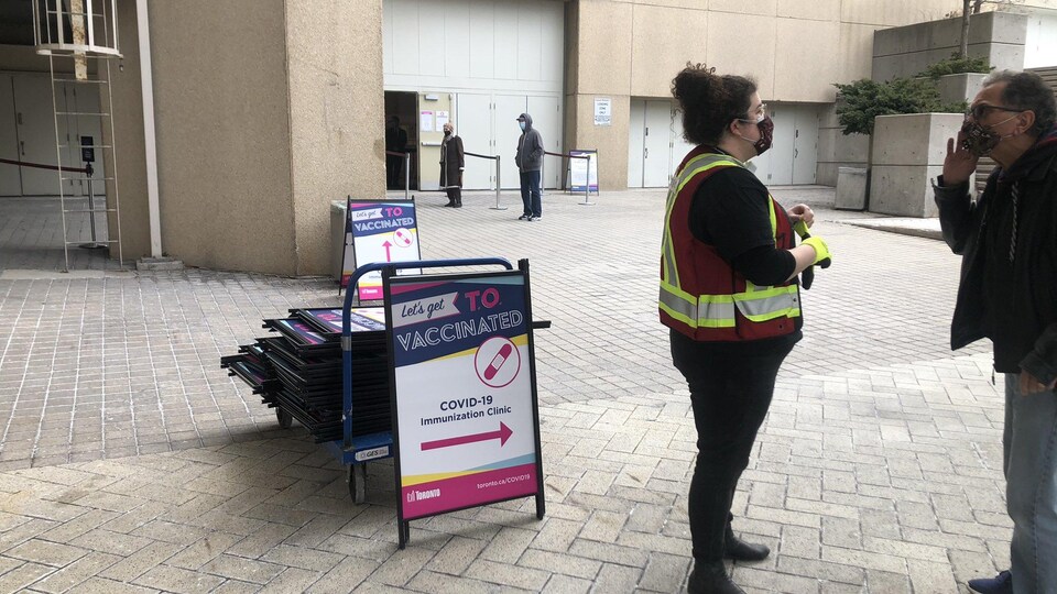 Une pancarte avec une flèche indique où se faire vacciner devant le Palais des congrès. Une dame avec un masque à l'extérieur s'entretient avec un possible patient, lui aussi avec un masque.