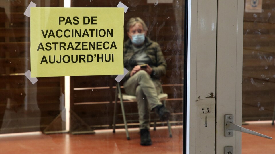 « Pas de vaccination AstraZeneca aujourd'hui », peut-on lire sur une affiche apposée sur porte vitrée. En arrière-plan, un homme masqué est assis.