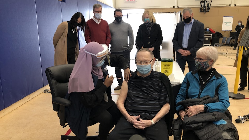 Un homme âgé reçoit un vaccin, derrière lui, le maire et quatre autres personnes.