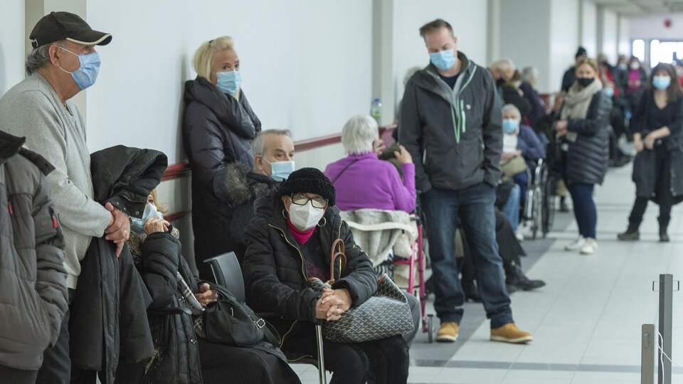 Des dizaines de personnes patientent en file. Certaines sont assises; d'autres se tiennent debout le long d'un mur. Elles portent toutes un couvre-visage.
