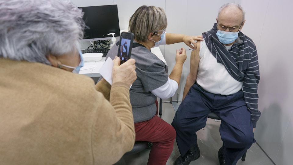 L'homme est assis sur une chaise et se fait vacciner. Sa femme prend une photo avec son cellulaire.