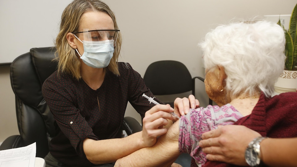 Une femme portant un masque et une visière administre un vaccin à une femme âgée. 