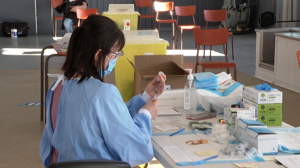 Une infirmière prépare une dose de vaccin. Des chaises sont disposées dans une grande salle pour les prochains patients.