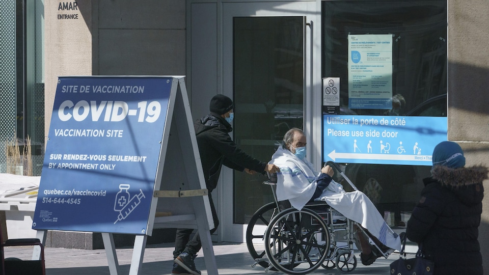 Une pancarte annonçant la vaccination devant l'hôpital; on aperçoit un patient en fauteuil roulant portant un masque, qu'un accompagnateur aide à se déplacer.