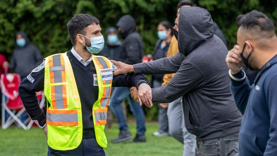Un homme attrape le bras d'un garde de sécurité dans un parc où l'on voit des gens faire la queue.