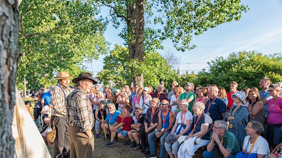 Une foule écoute deux comédiens en costume d'époque, sous les arbres.