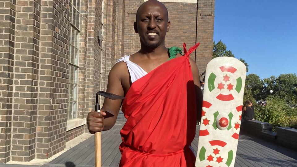 Valéry Mucowintore devant un tambour tenant une houe et un bouclier à l'effigie du drapeau burundais.