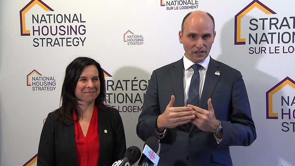Les deux politiciens sont debout devant un panneau sur lequel est écrit « Stratégie nationale sur le logement ». Valérie Plante sourit pendant que Jean-Yves Duclos donne des explications.