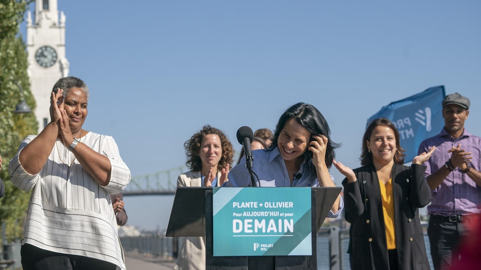 Valérie Plante sourit devant le micro, alors que ses candidats l'applaudissent, derrière.