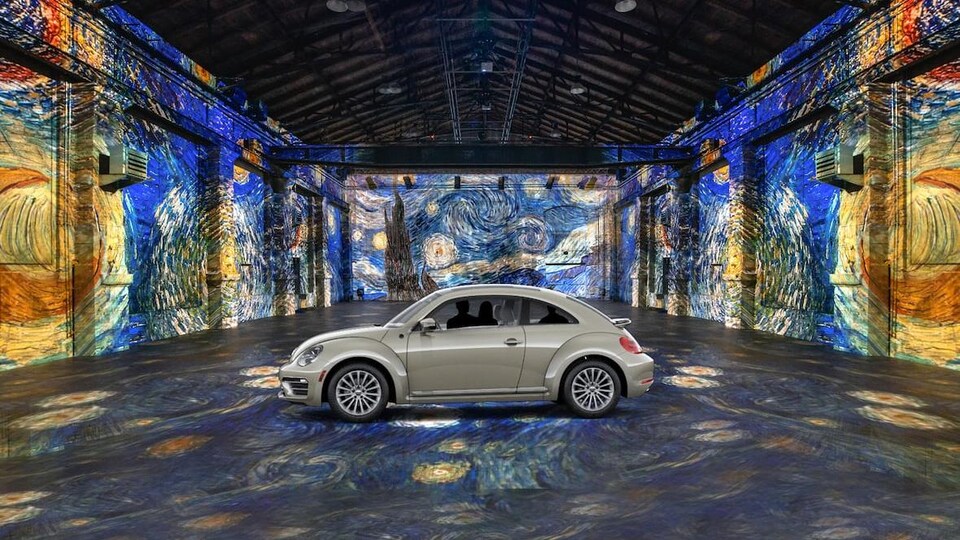 Une voiture au milieu d'un hangar désert. Des oeuvres de Van Gogh sont projetées sur le sol et sur les murs.