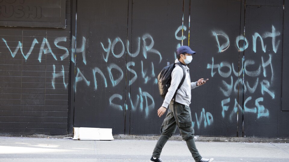 Un homme portant un masque passe devant un mur où on peut lire : « Lavez vos mains et ne touchez pas votre visage ».