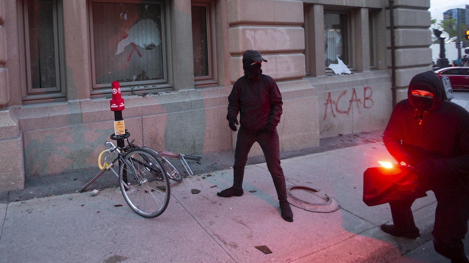 Deux hommes masqués vêtus de noir allument une fusée éclairante près d'une fenêtre brisée et d'un vélo vandalisé.
